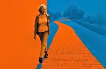 An older woman wearing glasses and a jacket walks confidently along a brightly colored, winding brick path that stretches into the distance, with an orange and blue color scheme dominating the image.