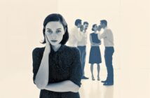 A woman in the foreground looks serious and isolated, while four people in the background stand in pairs, talking and whispering to each other. The scene suggests feelings of exclusion or loneliness.