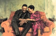 A joyful couple sits close together on an orange sofa, laughing and holding hands. The man wears a dark shirt and the woman wears a bright floral dress. They appear happy and relaxed in a cozy living room.