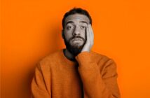 A man with a beard, wearing an orange sweater, rests his face on his hand and looks tired or bored. The background is bright orange, and the man's face is in grayscale.
