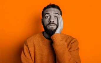 A man with a beard, wearing an orange sweater, rests his face on his hand and looks tired or bored. The background is bright orange, and the man's face is in grayscale.