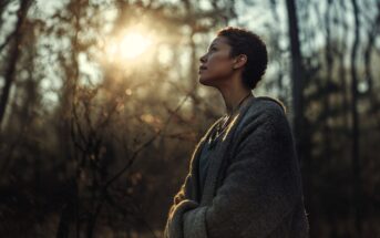 A person with short hair, wearing a cozy sweater, stands in a sunlit forest, looking thoughtfully upward as sunlight filters through the trees.