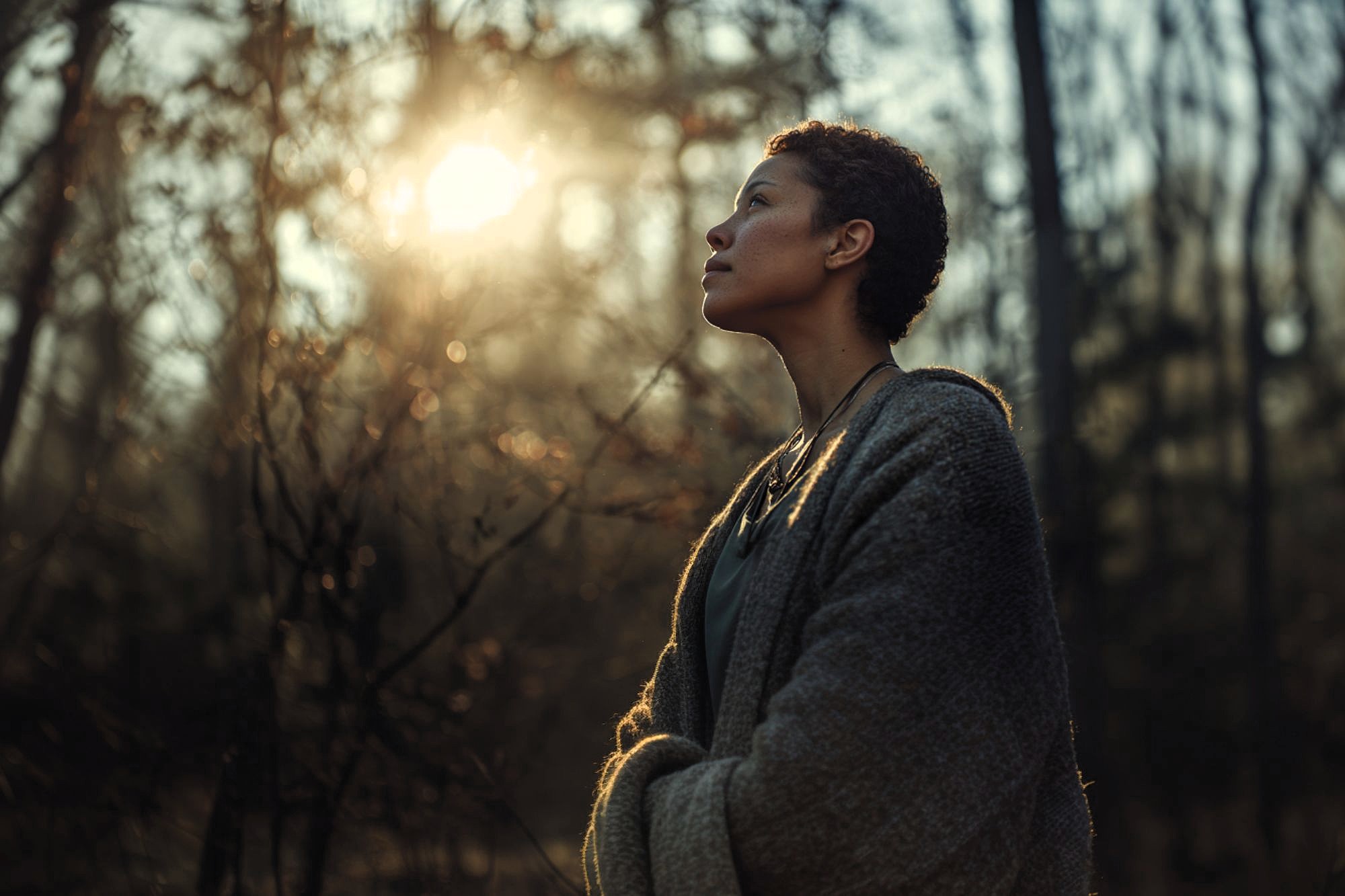 A person with short hair, wearing a cozy sweater, stands in a sunlit forest, looking thoughtfully upward as sunlight filters through the trees.