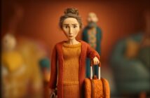 A claymation-style woman with gray hair in a bun, wearing an orange dress and cardigan, stands holding a suitcase. She looks concerned. Blurry figures and a couch are seen in the orange-toned background.