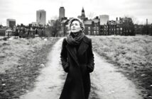 A woman in a long dark coat and scarf walks on a dirt path with her hands in her pockets, looking up. The background features city buildings and a cloudy sky. The image is black and white.