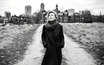 A woman in a long dark coat and scarf walks on a dirt path with her hands in her pockets, looking up. The background features city buildings and a cloudy sky. The image is black and white.