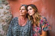 Two women, one older with short gray hair and one younger with long blonde hair, stand close together against a textured wall, both looking thoughtfully into the distance. They wear floral-patterned tops.