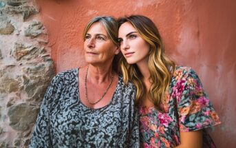 Two women, one older with short gray hair and one younger with long blonde hair, stand close together against a textured wall, both looking thoughtfully into the distance. They wear floral-patterned tops.