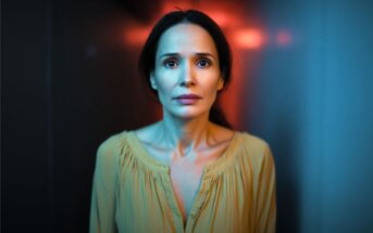 A woman with long brown hair wearing a yellow blouse stands in a dimly lit hallway, looking forward with a concerned expression. Blue and red lighting creates a dramatic atmosphere around her.