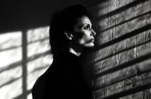 A person with short hair and a serious expression stands in profile against a textured wall, with dramatic shadows and light patterns from window blinds cast across their face and the wall.