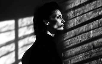 A person with short hair and a serious expression stands in profile against a textured wall, with dramatic shadows and light patterns from window blinds cast across their face and the wall.