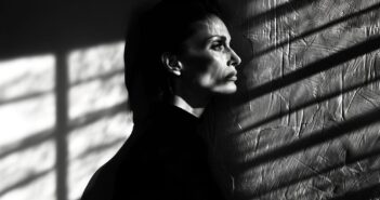 A person with short hair and a serious expression stands in profile against a textured wall, with dramatic shadows and light patterns from window blinds cast across their face and the wall.