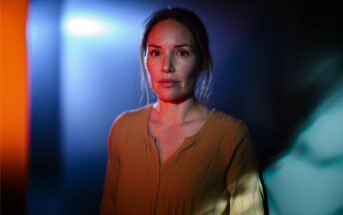 A woman in a yellow blouse stands in dramatic lighting with blue and orange tones, looking forward with a serious expression.