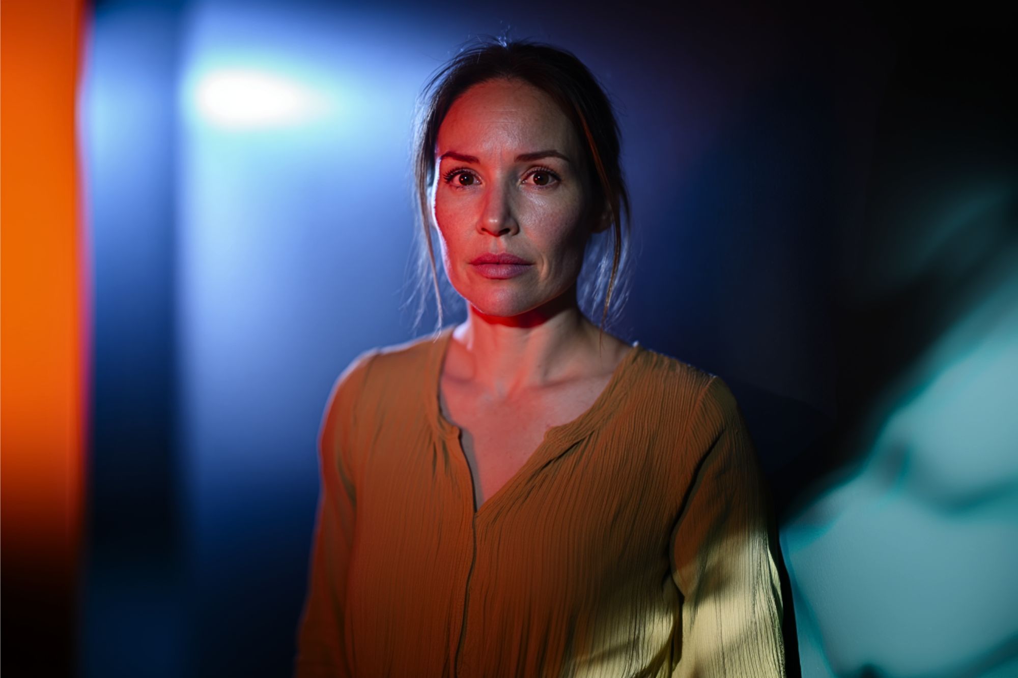 A woman in a yellow blouse stands in dramatic lighting with blue and orange tones, looking forward with a serious expression.