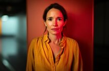 A woman with brown hair in a yellow blouse stands against an orange wall, looking directly at the camera with a serious, slightly worried expression. Red light casts a dramatic glow on her and the background.