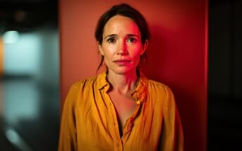 A woman with brown hair in a yellow blouse stands against an orange wall, looking directly at the camera with a serious, slightly worried expression. Red light casts a dramatic glow on her and the background.