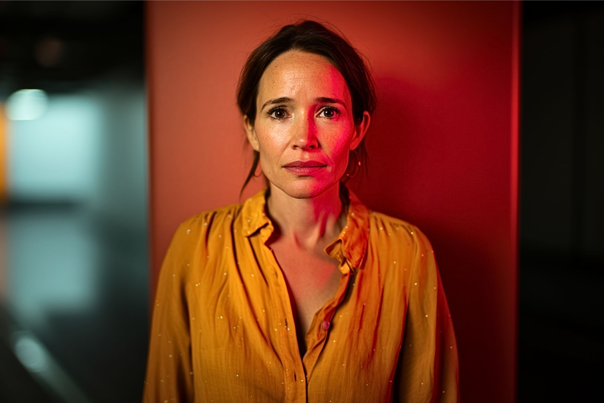 A woman with brown hair in a yellow blouse stands against an orange wall, looking directly at the camera with a serious, slightly worried expression. Red light casts a dramatic glow on her and the background.