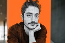 A stylized portrait of a person with curly hair and a mustache, resting their chin on their hand, set against a bright orange background with blurred indoor surroundings.