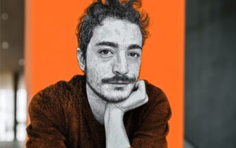A stylized portrait of a person with curly hair and a mustache, resting their chin on their hand, set against a bright orange background with blurred indoor surroundings.
