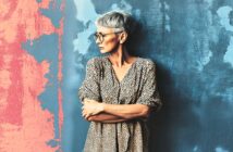 Older woman with short gray hair and glasses stands against a textured wall painted blue and pink, looking to her left with arms folded, wearing a spotted, loose-fitting dress.