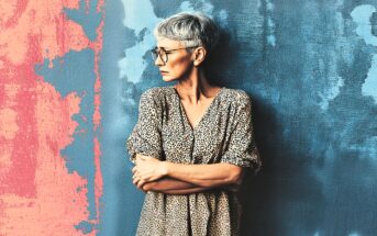 Older woman with short gray hair and glasses stands against a textured wall painted blue and pink, looking to her left with arms folded, wearing a spotted, loose-fitting dress.