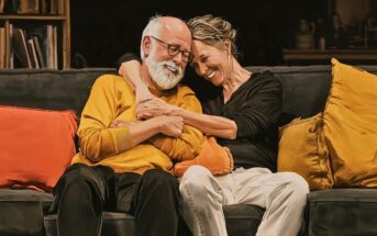 An older couple sitting on a couch, embracing and smiling with their heads resting against each other.