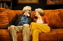 An older couple with gray hair and glasses sit close together on an orange sofa, smiling and laughing warmly at each other in a cozy living room setting.