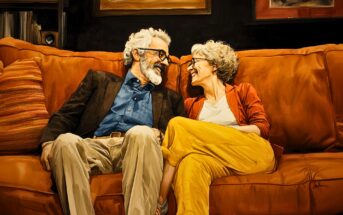 An older couple with gray hair and glasses sit close together on an orange sofa, smiling and laughing warmly at each other in a cozy living room setting.