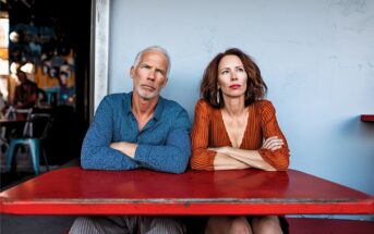 A mature man and woman sit at a red table with their arms crossed, looking serious. The man wears a blue shirt, and the woman wears an orange dress. They are seated outside against a pale wall.