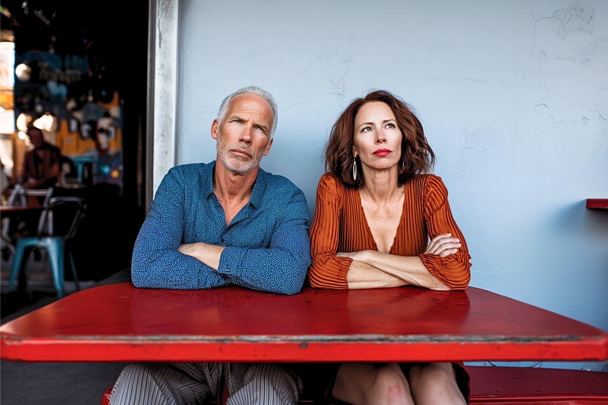 A mature man and woman sit at a red table with their arms crossed, looking serious. The man wears a blue shirt, and the woman wears an orange dress. They are seated outside against a pale wall.