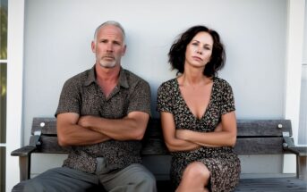 A middle-aged man and woman sit on a bench with arms crossed, looking serious. Both wear casual, patterned clothing. The background is a plain, light-colored wall.