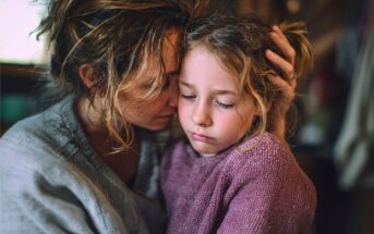 A woman gently hugs a sad young girl, comforting her. Both have messy hair and wear cozy sweaters. The scene is warm and intimate, suggesting care and support during a difficult moment.