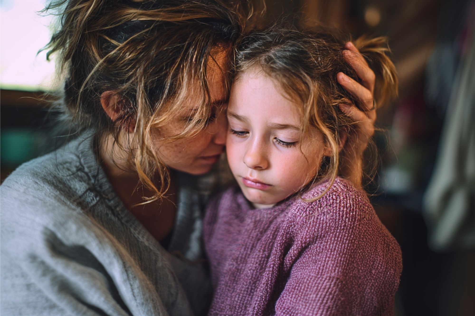 A woman gently hugs a sad young girl, comforting her. Both have messy hair and wear cozy sweaters. The scene is warm and intimate, suggesting care and support during a difficult moment.
