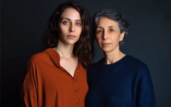 Two women stand close together against a dark background. One has long, wavy brown hair and wears a rust-colored shirt. The other has short, gray hair and wears a dark blue sweater. Both look calmly at the camera.