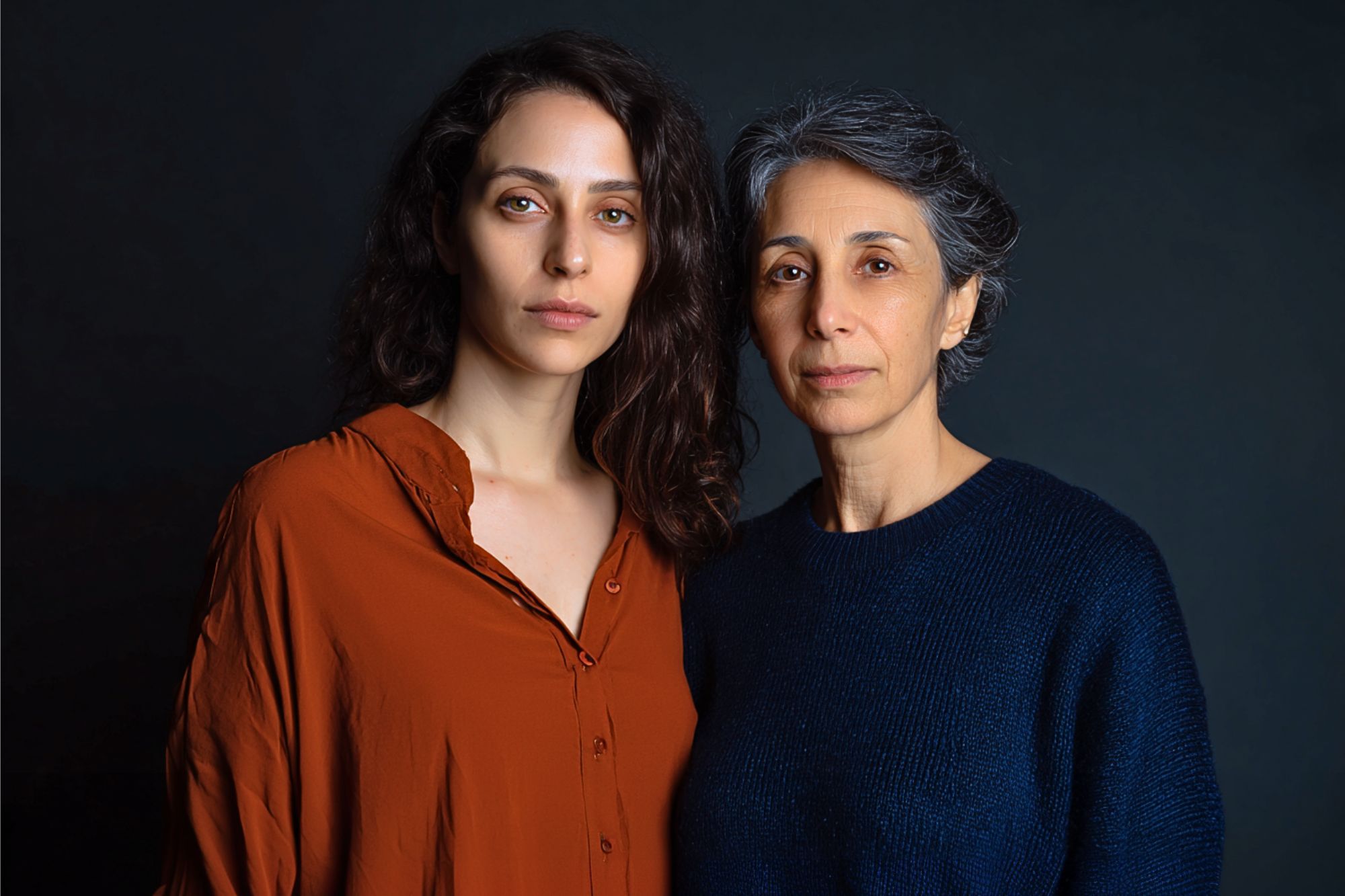 Two women stand close together against a dark background. One has long, wavy brown hair and wears a rust-colored shirt. The other has short, gray hair and wears a dark blue sweater. Both look calmly at the camera.