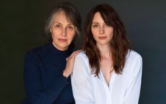 An older woman with graying hair and a younger woman with brown hair pose together, with the older woman resting a hand on the younger woman’s shoulder.