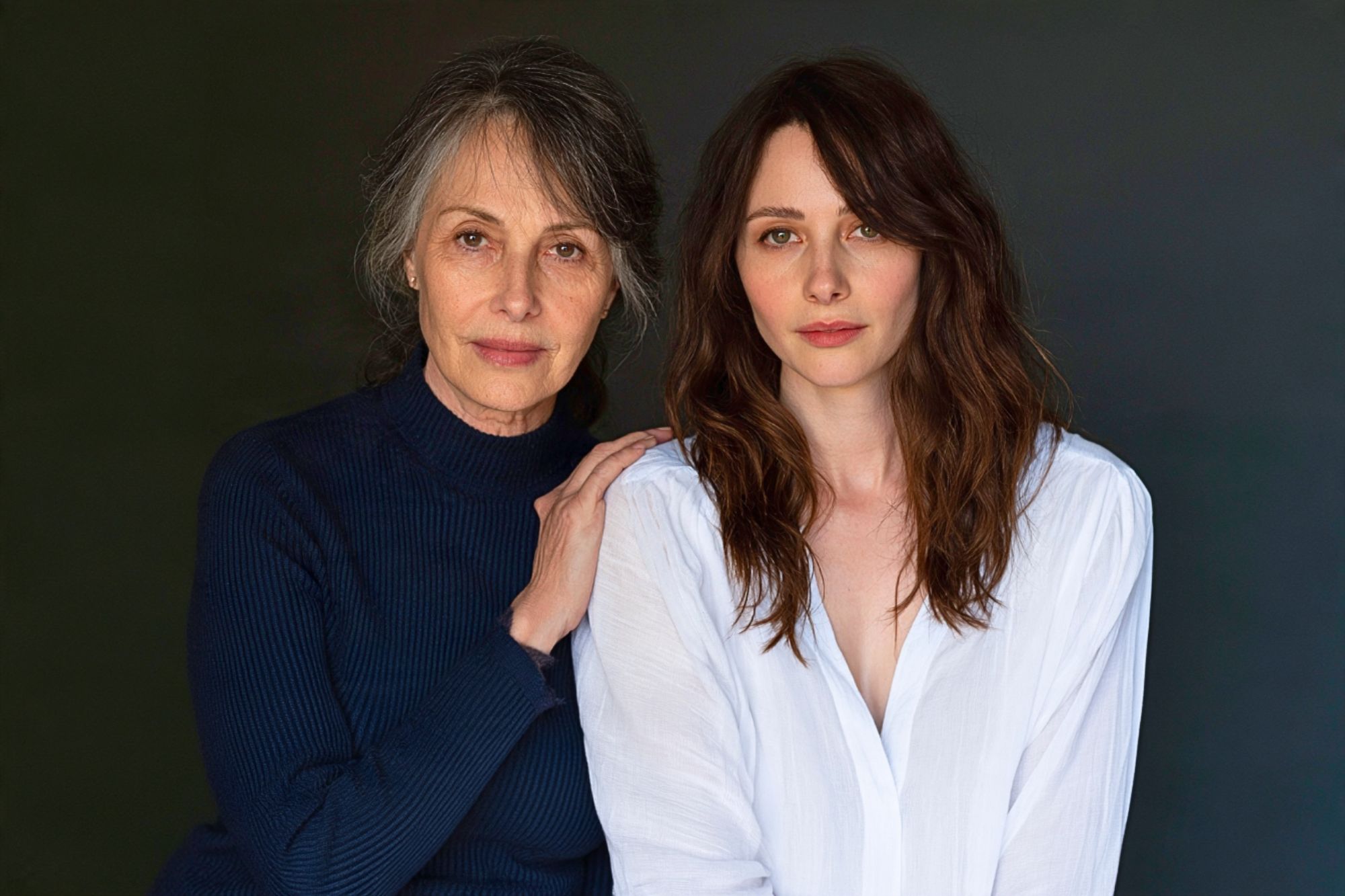 An older woman with graying hair and a younger woman with brown hair pose together, with the older woman resting a hand on the younger woman’s shoulder.