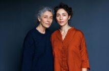 Two women stand close together against a dark blue background. The older woman has gray hair and wears a navy sweater, while the younger woman has curly dark hair and wears a loose orange shirt. They look calmly at the camera.