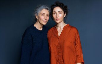 Two women stand close together against a dark blue background. The older woman has gray hair and wears a navy sweater, while the younger woman has curly dark hair and wears a loose orange shirt. They look calmly at the camera.