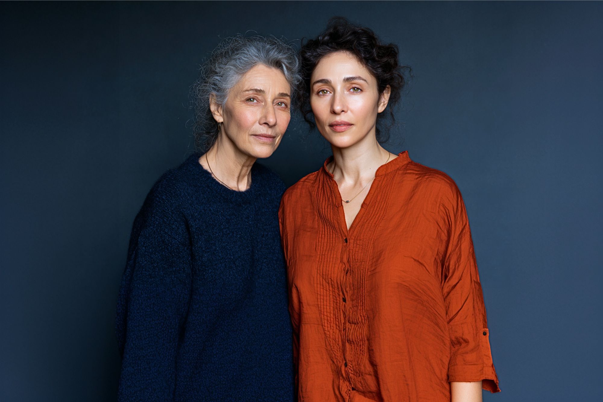 Two women stand close together against a dark blue background. The older woman has gray hair and wears a navy sweater, while the younger woman has curly dark hair and wears a loose orange shirt. They look calmly at the camera.