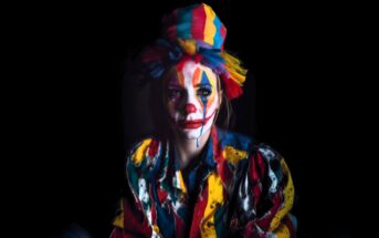 A person dressed as a clown with colorful makeup, a rainbow-striped hat, and a vibrant, multicolored outfit sits against a dark background. The clown has smeared face paint and a somber expression.