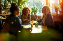 Three women sit at a sunlit café table, engaged in a lively conversation. Sunlight streams through the window, and green plants surround them, creating a warm and inviting atmosphere.