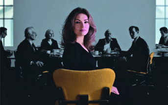 A woman in a black outfit sits on a yellow chair facing the camera, with several people blurred in the background around a conference table in a dimly lit room.