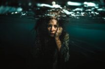 A woman with long hair and a distressed expression is submerged underwater, with her face partially visible above the surface. One hand rests on her cheek as light filters through the rippling water.