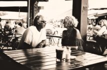 A man and woman sit across from each other at an outdoor café table, engaged in conversation. The café is busy, with other patrons in the background. The image is in black and white.