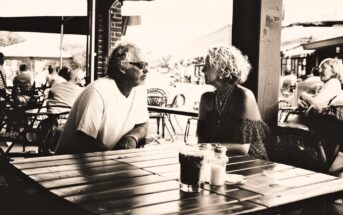 A man and woman sit across from each other at an outdoor café table, engaged in conversation. The café is busy, with other patrons in the background. The image is in black and white.