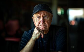 An elderly man wearing a black cap and dark shirt sits with his hand resting on his chin, looking thoughtfully at the camera. The background is blurred, drawing focus to his expressive face and contemplative expression.