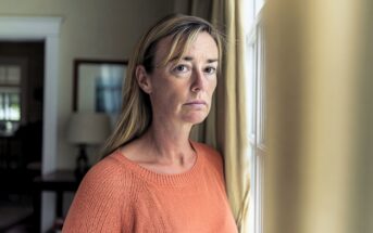 A woman with long blonde hair and an orange sweater stands near a window, looking toward the camera.