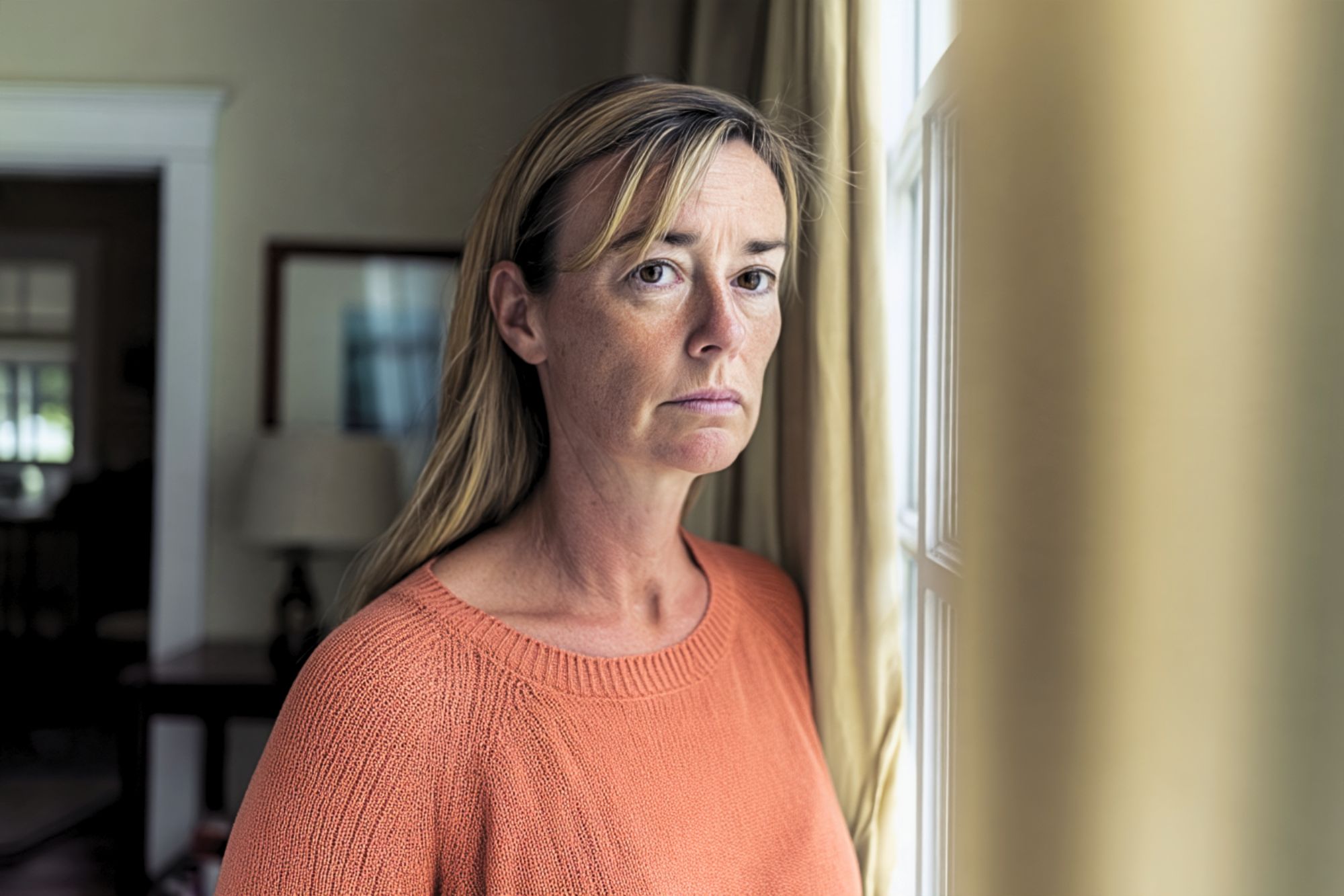 A woman with long blonde hair and an orange sweater stands near a window, looking toward the camera.