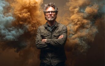 A man with gray hair and glasses stands confidently with arms crossed, wearing a casual jacket. Dramatic orange and blue smoke billows around him against a dark background.